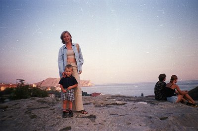 A woman and young boy stand on a rocky outcrop, overlooking a seaside landscape with a distant town and coastline. The woman ...