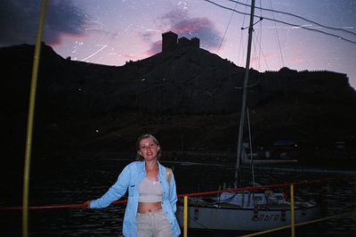 A woman stands on a boat deck, posed with a dramatic cliffside fortress illuminated by a pink sunset. The fortress is likely ...