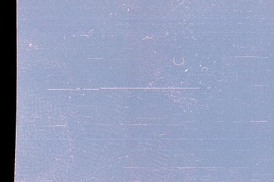 Close-up of a heavily distressed surface, exhibiting extensive scratches and dust particles across a pale blue field. The tex...