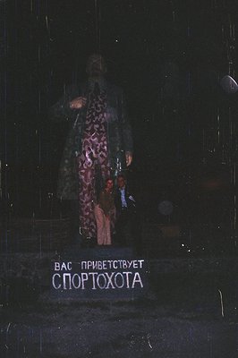 A man in a patterned coat and dark pants stands before a neon sign reading "Спортохота" (Sportohota) in Cyrillic script. The ...