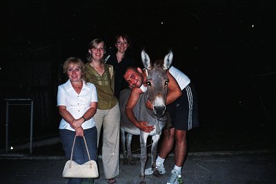 Four individuals pose outdoors at night: three women and one man embracing a donkey. The man wears a white tank top, and the ...