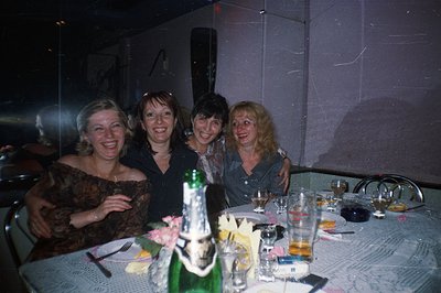 Four women share a celebratory moment at a table covered with a lace tablecloth, bubbly champagne, and glassware. The image s...