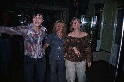 Three women stand close together, laughing, in what appears to be a dimly lit exterior space. They are wearing patterned shir...