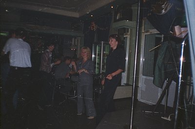 A group gathers in a dimly-lit, possibly nightclub setting. Two women in flared trousers, one blonde, stand near a mirrored b...