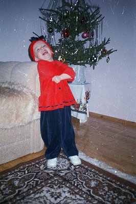 Young child in a festive red velvet outfit, including hat, poses joyfully near a decorated Christmas tree. Visible grain sugg...
