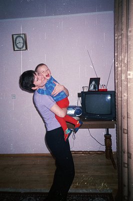A young woman playfully tosses a toddler into the air indoors. The scene includes a vintage wood TV console and a decorative ...
