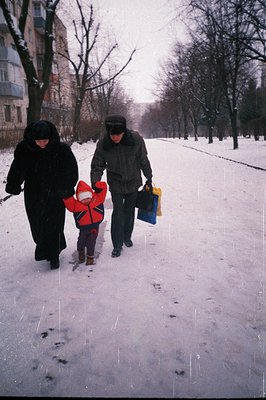 A man and child walk along a snowy, tree-lined street. The man carries shopping bags, while the child wears a bright red snow...