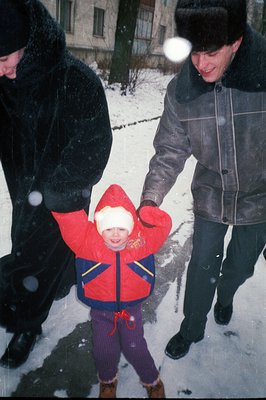 A young child in a red snowsuit stands with their arms outstretched, appearing to be playfully restrained by an adult. The sc...