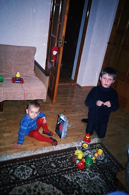 Two young boys sit amidst toys on a patterned rug. One wears a blue plaid shirt and red pants, the other a dark sweater. Like...