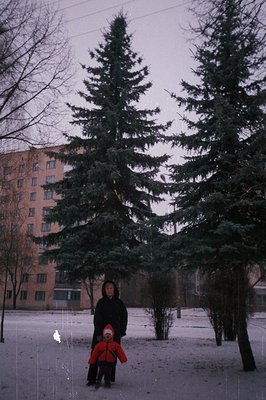 A snowy courtyard features a mother and child in winter coats amidst evergreen trees. Behind them, a Soviet-era apartment blo...