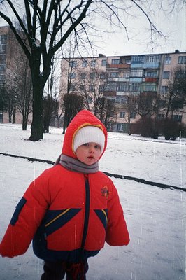 A toddler bundled in a vibrant red winter coat and hood stands in a snow-covered courtyard. A large apartment building, typic...