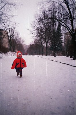 A child in a vibrant red snowsuit runs through a snow-covered park. Bare trees line the background, hinting at a cold, possib...