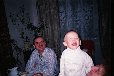 A smiling man sits next to a laughing toddler wearing a cream-colored turtleneck sweater, seemingly interacting playfully. In...