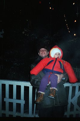 A man carries a toddler on his shoulders, both bundled in winter gear. The man wears a dark jacket; the child is in a vibrant...