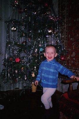 A young boy stands proudly before a Christmas tree adorned with tinsel and ornaments. He wears a blue plaid shirt and white p...