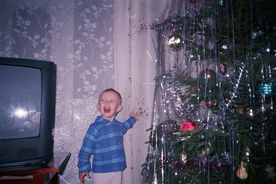 A young boy with a wide-open mouth stands near a brightly decorated Christmas tree. The scene features a vintage television s...