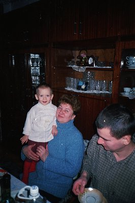 A young boy stands on the lap of a woman in a blue sweater, while a man holds them from the side in what appears to be a dini...