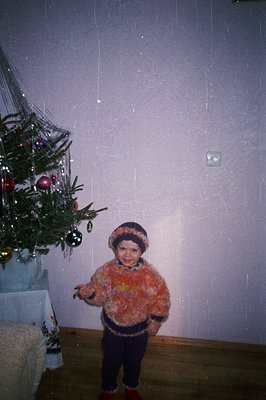 A young child stands near a decorated Christmas tree, wearing a textured orange sweater, dark pants, and a fur-trimmed hat. T...