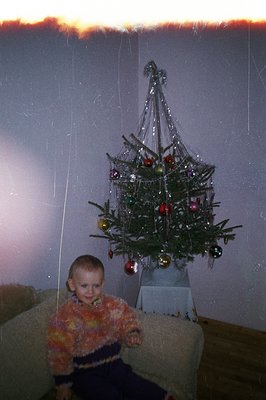 A young child with short hair sits beside a unique, sputnik-style aluminum Christmas tree. The tree, decorated with glass orn...