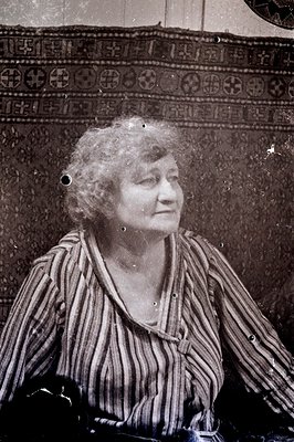 A portrait of a woman with tightly curled hair, seated and looking toward the camera. She wears a striped, button-down blouse...