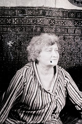 A monochrome portrait captures a woman with curly gray hair and a noticeable overbite, seated and gazing slightly to the side...