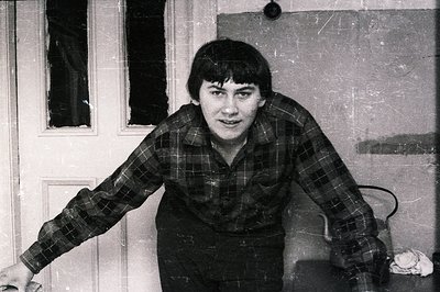 A man with a short, dark haircut stands facing the camera, arms outstretched near a weathered door. He wears a plaid shirt an...