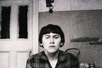 B&W portrait of a young man with short, dark hair, seated. He wears a plaid shirt and exhibits a slight frown. The background...