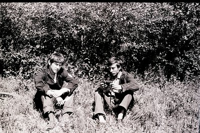 Two young men sit in tall grass, appearing contemplative. One holds a small object, perhaps a camera or tool. Clothing sugges...