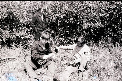 Three young men relax in overgrown grass with a bicycle partially visible. Two are seated, apparently examining a small objec...