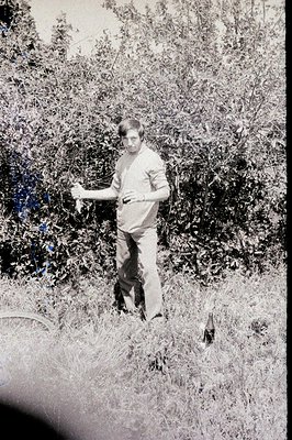 A young man in a long-sleeved sweater and trousers stands amidst dense foliage, holding a small object. Appears to be a candi...