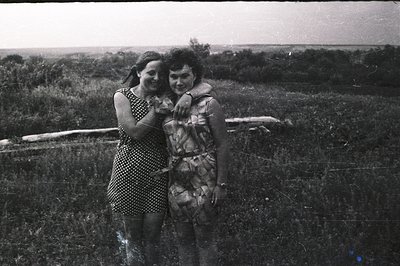 Two young women in patterned dresses stand in a field with a treeline backdrop. A fallen log sits prominently in the foregrou...