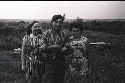 Three individuals posed outdoors, likely in a rural setting with tall grasses and a distant horizon. The man stands center, a...