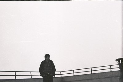 A figure stands silhouetted on a rooftop, overlooking a hazy sky. Concrete railings define the edge of the space. Appears to ...