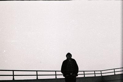 A solitary figure stands at a railing overlooking an expanse of white space, likely a building facade. The subject is silhoue...
