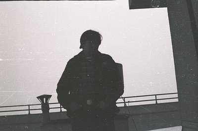 A shadowed figure stands within an architectural structure, likely a balcony or observation deck. The composition emphasizes ...