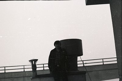 A young man stands near a large cylindrical structure, possibly industrial or architectural. Visible railing suggests a rooft...