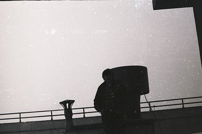 A silhouetted figure stands on a rooftop, framed by a modern railing and architectural elements. The stark contrast emphasize...