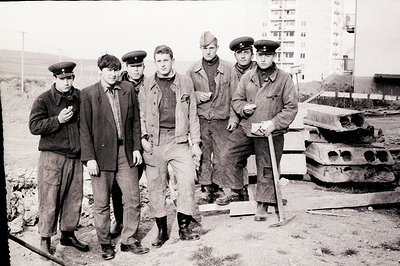 Six young men in work clothes and caps pose outdoors, standing near heavy machinery. Likely a construction or infrastructure ...