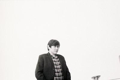 A young man in a dark blazer and plaid shirt gazes slightly off-camera. The composition is minimalist, set against a plain, l...