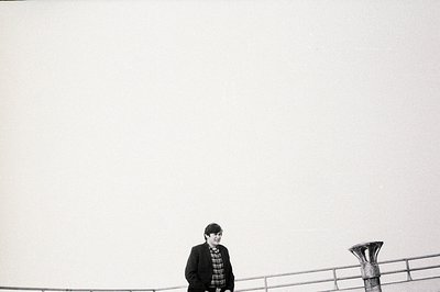 A man in a plaid shirt stands near a railing, facing left, against a stark, overcast sky. The composition emphasizes vastness...