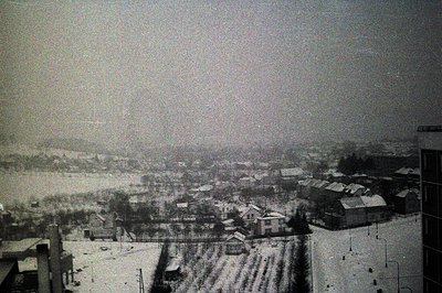 A monochrome, high-angle view captures a snow-covered townscape. Buildings with pitched roofs are densely packed, partially o...