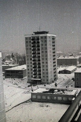Tall, austere apartment block dominates a snow-covered urban landscape. Visible balconies suggest a 1960s-70s Soviet-era cons...