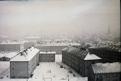 Overcast view of a densely populated urban area blanketed in snow. Multi-story apartment buildings dominate the scene, featur...