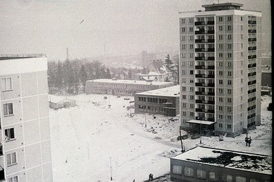 A stark, snowy cityscape featuring brutalist apartment blocks, likely from the 1960s-70s. The tall building showcases repetit...