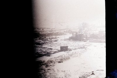 Aerial view of a snow-covered industrial area. Several low-rise buildings and rail lines are visible under a heavy snowfall. ...