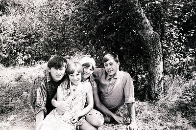 Four young people pose for a portrait amidst lush foliage, likely a wooded area. The subjects display 1960s or 70s fashion: f...