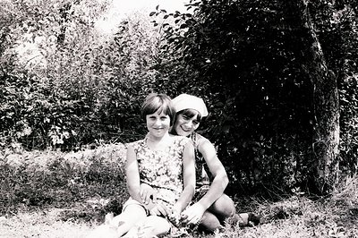 Two young girls sit on grass, partially obscured by foliage. One wears a floral dress and the other, a cap. Appears to be a c...