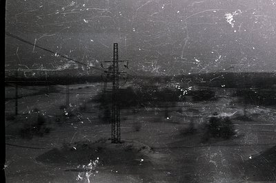A stark, monochrome landscape featuring an electrical pylon dominating the midground. Sparse, low vegetation and a distant, h...