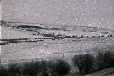 Hilly, undulated landscape with scattered trees & distant buildings. Appears to be rural terrain, possibly farmland. Visible ...