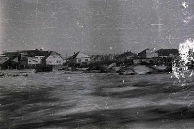 A view of seaside homes threatened by coastal erosion. Buildings with tiled roofs cling to a slope above a turbulent sea. Vis...